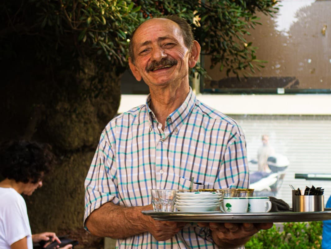 An elderly Italian waiter smiling as he serves coffee at outdoor tables